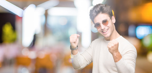 Young handsome man wearing sunglasses over isolated background very happy and excited doing winner gesture with arms raised, smiling and screaming for success. Celebration concept.