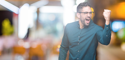 Young handsome business man wearing glasses over isolated background angry and mad raising fist frustrated and furious while shouting with anger. Rage and aggressive concept.