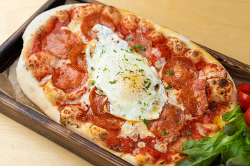 Classic rome pizza with crust, tomato sauce, spicy pepperoni and fried egg in the centre, served on wooden tray