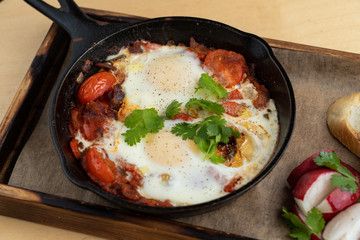 Traditional israeli breakfast: shakshuka with tomatoes, salami, chili pepper and fried eggs, served in hot cast pan with toasts and radish