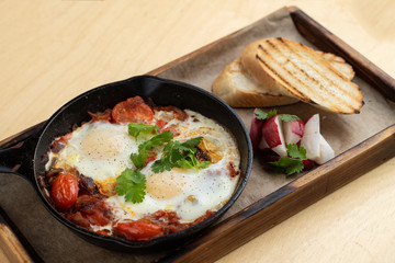 Traditional israeli breakfast: shakshuka with tomatoes, salami, chili pepper and fried eggs, served in hot cast pan with toasts and radish