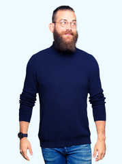 Young blond man wearing glasses and turtleneck sweater smiling looking side and staring away thinking.