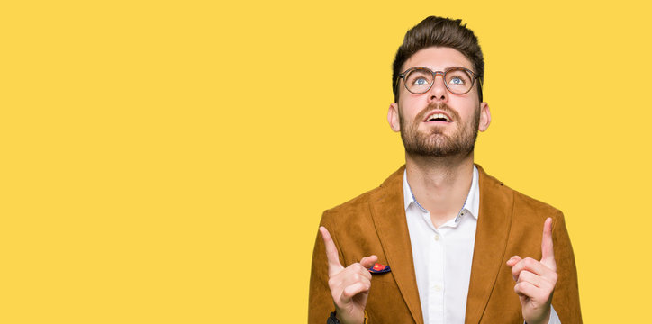 Young handsome business man wearing glasses amazed and surprised looking up and pointing with fingers and raised arms.