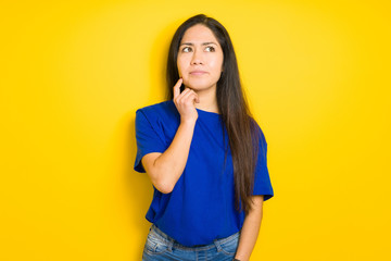 Beautiful brunette woman wearing blue t-shirt over yellow isolated background with hand on chin thinking about question, pensive expression. Smiling with thoughtful face. Doubt concept.