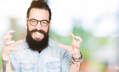 Young hipster man with long hair and beard wearing glasses Shouting frustrated with rage, hands...