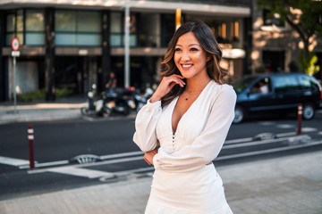 Fototapeta premium Summer sunny lifestyle portrait of young stylish asian girl on the street of big modern city. She wears white dress