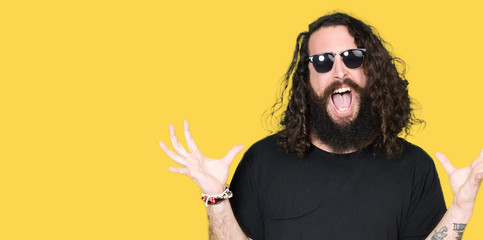 Young hipster man with long hair and beard wearing sunglasses crazy and mad shouting and yelling...