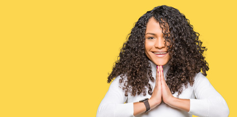 Young beautiful woman with curly hair wearing turtleneck sweater praying with hands together asking...