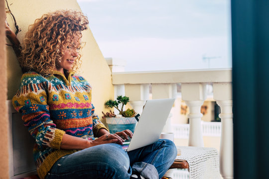 Beautiful adult young curly woman at work on the terrace at home with laptop computer - coloured hand made stylisand trendy sweater for autumn or winter season. concept