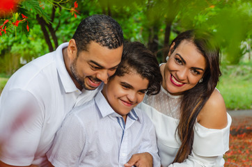 Obraz premium couple of Latin American men and women, with boy suffers autism, happy in a portrait family outdoors together in a park, the three laughing hugging