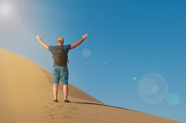 Fototapeta na wymiar A man with backpack on the sand dune happy to be there. Male rise his hands in desert landscape