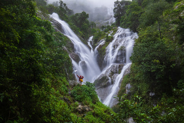 Fototapeta na wymiar The girl in red sweater touring on Pi-tu-gro waterfall, Beautiful waterfall in Tak province, ThaiLand.