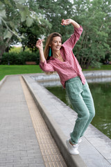 young European girl, natural appearance, long hair, beautiful face, green pants, pink shirt, white sneakers. To walk to pose on path near lake in city park. Stylish linen womens clothing.