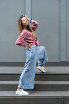 young Caucasian girl, natural appearance, long hair, beautiful face, blue wide pants, crimson color shirt, white sneakers actively pose gray wall background. Comfortable linen womens clothing.