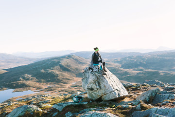 Athletic female hiker rests on boulder and looks out at view of Norwegian landscape with lakes and...