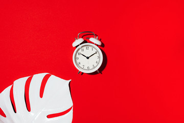White alarm clock and monstera leaf over red background with copy space. Top view. Flat lay. Wake up alert concept. Morning routine.