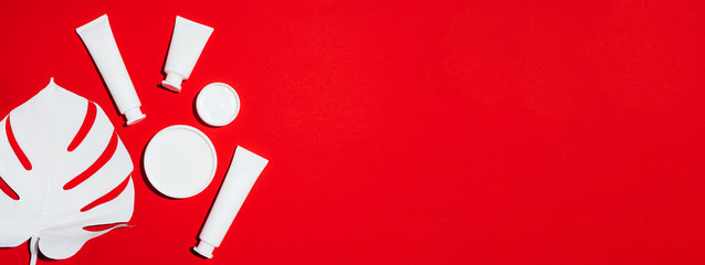 Cosmetics, skin care, beauty, body treatment concept. White cosmetic jar, tube, bottle and tropical monstera leaf over red background. Top view. Flat lay. Mock-up.