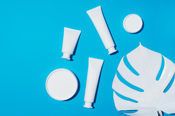 White plastic cosmetic jar, tube, bottle and tropical monstera leaf over blue background. Top view. Flat lay. Mock-up. Cosmetics, skin care, beauty, body treatment concept