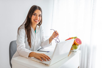 Portrait of young smiling female nutritionist in the consultation room. Portrait of beautiful smiling nutritionist looking at camera and showing healthy vegetables in the consultation.