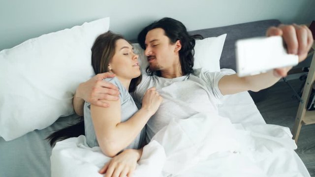 Man and woman taking selfie in bed at home using smartphone camera having fun showing thumbs-up and smiling. People, modern technology and photo concept.