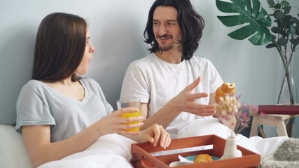 Happy young people are laughing chatting eating breakfast in bed at home enjoying croissants and orange juice. Youth lifestyle, relationship and food concept. - Powered by Adobe