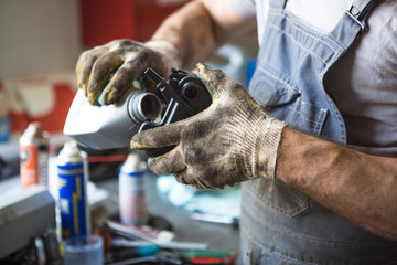 A mechanic in dirty gloves and overalls pours engine oil from a canister into the engine. The...