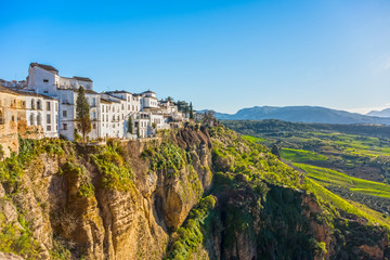 Ronda, Spain: Landscape of white houses on the green edges of steep cliffs with mountains in the background. - obrazy, fototapety, plakaty