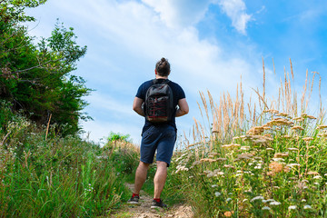 Hiker in blue shorts standing on the mountain trail, almost at the top, with backpack on his back, surrounded with green grass and field flowers, on a sunny day with bright blue sky