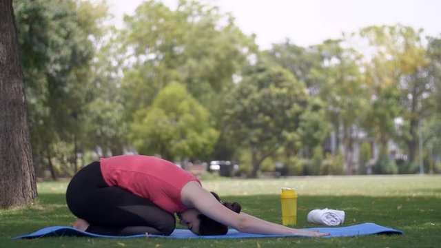 Young attractive woman practicing Balasana (child pose) on a yoga mat - healthy lifestyle. Calm Indian lady doing child pose under a tree while sitting on mat in sportswear to be fit and healthy