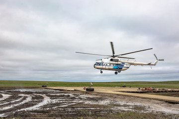 A polar aviation helicopter is preparing to land on a remote heliport in the northern tundra, not far from a drilling oil well. Wooden platform with lying equipment. Gloomy northern sky. The arrival o