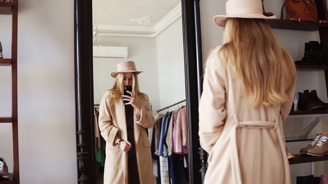 Attractive blonde girl standing in front the mirror in beige coat and trendy hat, making a photo of look with her smartphone. Footage from the backside