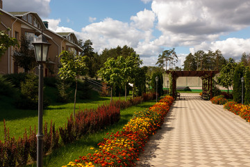 Fototapeta na wymiar A quiet street in a modern country village on a sunny summer day. In the foreground Street lamp. The paved path is laid past flower beds, ornamental shrubs and trees.