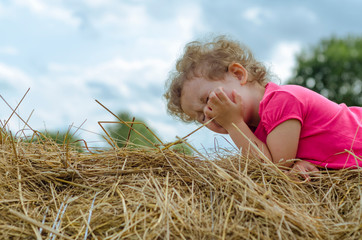 Naklejka na ściany i meble the little girl closes her eyes and squints from the sun. German child lies on hay bale during Oktoberfest. soft focus.