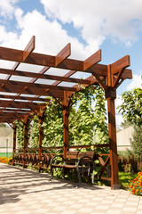 Wooden pergollas from natural wood with comfortable benches in a modern country village on a sunny summer day. The paved path is laid past flower beds, ornamental shrubs and trees. Russia