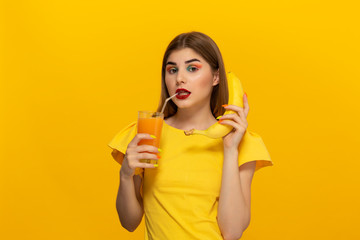 Close-up portrait of a beautiful young girl holding glass of ofange juice and banana close to face isolated over yellow background.