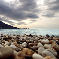 Obraz premium Calm water at coast. Ripple of water near shoreline. Silhouette of city on skyline. Stone seashore before storm. Rocky beach near the bay.