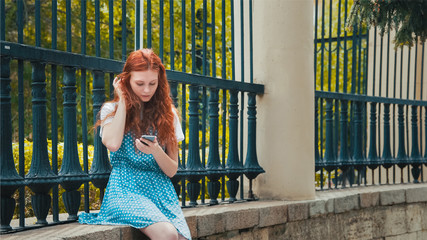 Ginger girl looks into the phone. Beauty redhead woman  reading. Red-haired freckled teenager sitting in park. Portrait with copyspace.
