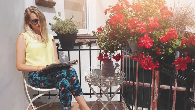 young attractive woman with sunglasses enjoying sun and reading magazine on romantic balcony with flower boxes