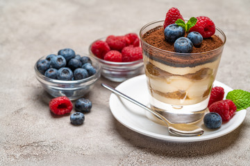 Classic tiramisu dessert with blueberries and raspberries in a glass and bowls with berries on concrete background