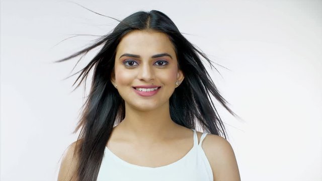 Closeup shot of a young beautiful girl giving a toothy smile against white background - lifestyle concept. Attractive Indian woman with long blowing hair  happily smiling while looking towards the ...