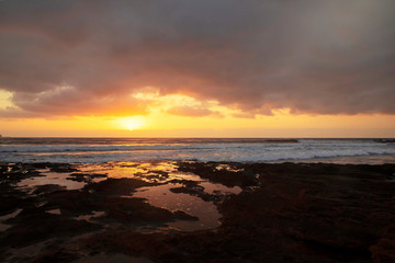 Glorious sunrise reflected in the natural rock pools formed in the limestone shores of El Medano resort, Tenerife, Canary Islands, Spain, vibrant seascape with overcast sky and golden haze light