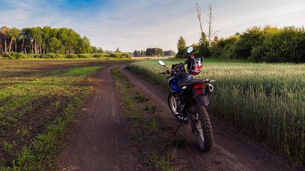 Obraz premium Tourist motorcycle with red helmet on the handlebar stands on picturesque country road, next to green field of wheat