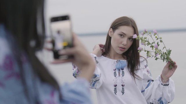 Portrait of attractive young brunette girl in long embroidered dress holding flower near face posing while her friend taking photo of her. Concept of fashion, connection with nature, rural life