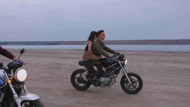 Young couple riding on vintage motorcycles during cloudy sunset on beach, slow motion