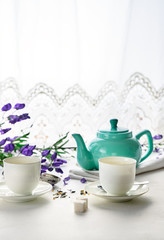 Two white tea cups with green tea, teal colored tea pot, purple flowers on white background with copy space