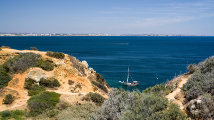 Vacation in Algarve - View of a a sailboat motoring on Atlantic near the coastline seen from above. Dry red soil cliffs and green vegetation in foreground, buildings in distance, blue sky white clouds