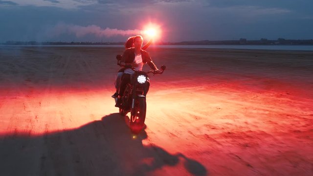 Couple riding on vintage motorcycle with red burning signal fire after sunset on beach, slow motion
