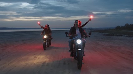 Two young couples riding on vintage motorcycles with red burning signal flares after sunset on beach, slow motion