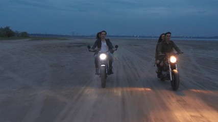 Cinematic shot of two young couples riding on vintage motorcycles after sunset on beach, slow motion