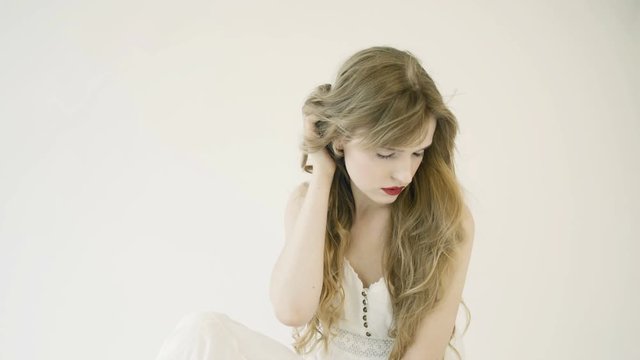 Girl with long hair in white long dress sits on white background in studio. Delicate facial features, nude makeup. Wind blows hair. She touches her hair, straightens her hair. 